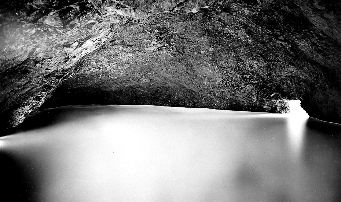 Fotografia dell'interno della sorgente Peschiera, con l'acqua che scorre silenziosa sotto una volta rocciosa naturale. L'immagine, ripresa con lunga esposizione, restituisce la luminosità e la purezza dell'acqua alla sua origine. Anno 1908.