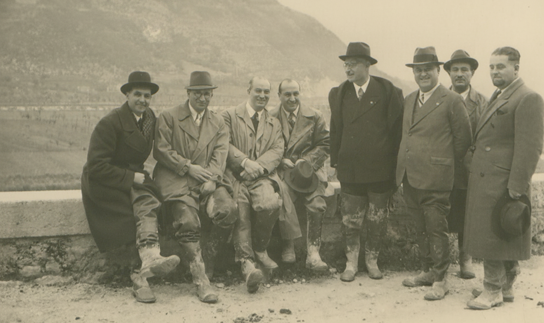 Gruppo di otto uomini in abiti formali e cappello, alcuni seduti su un muretto e alcuni in piedi, durante un sopralluogo ai lavori dell'Acquedotto Peschiera. Sullo sfondo, un paesaggio collinare. Anno 1938.