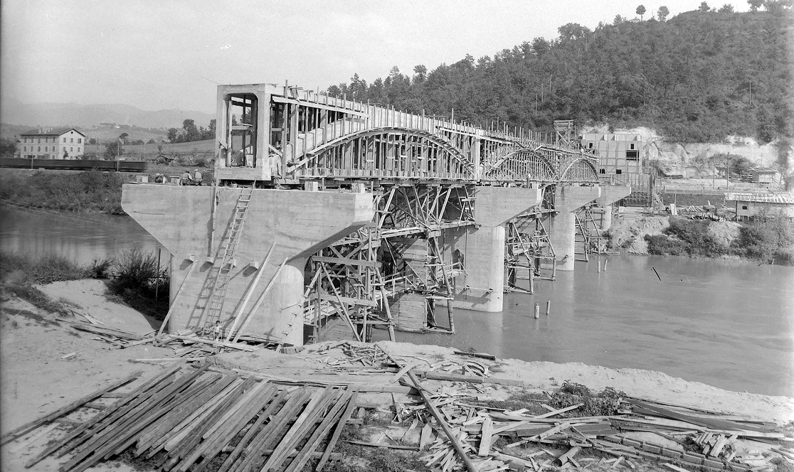 Costruzione di un ponte sull'Acquedotto Peschiera, 1940. L'immagine mostra le pile in cemento armato e la struttura ad arco in fase di assemblaggio, con le impalcature in legno ancora in opera e materiali da costruzione sul greto del fiume.