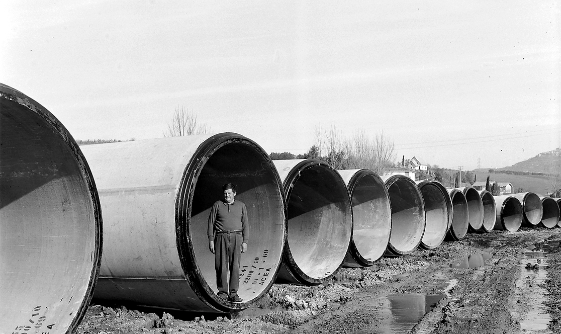 Un operaio ACEA in piedi all'interno di uno dei grandi tubi in cemento armato destinati all'ampliamento dell'Acquedotto Peschiera, per dare la misura delle dimensioni. Sul tubo è visibile la scritta "ACEA 69". Anni 1966–1971.