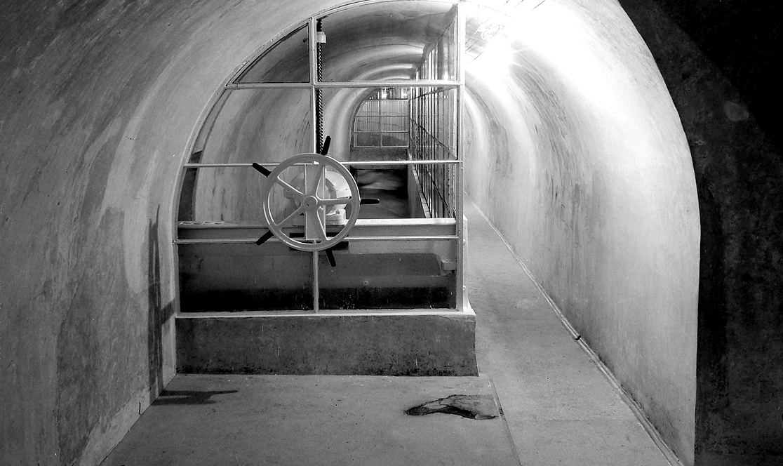 Interno di una galleria dell'Acquedotto Peschiera con volta a botte in muratura. Al centro dell'inquadratura, una valvola a volantino e una condotta metallica che si estende verso il fondo illuminato del tunnel. Anno 1975.