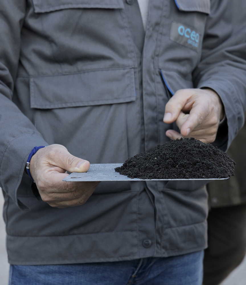 Dipendente Acea in abbigliamento da lavoro tiene in mano una paletta metallicacon un mucchietto di terriccio scuro, che indica con un dico
